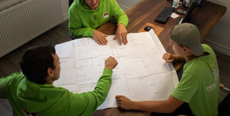 Drei Mitarbeiter sitzen an einem Tisch auf dem ein Grundriss eines Bauvorhabens liegt. Ein Mitarbeiter zeigt mit den Fingern auf einem bestimmten Bereich.