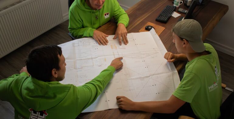 Drei Mitarbeiter sitzen an einem Tisch auf dem ein Grundriss eines Bauvorhabens liegt. Ein Mitarbeiter zeigt mit den Fingern auf einem bestimmten Bereich.