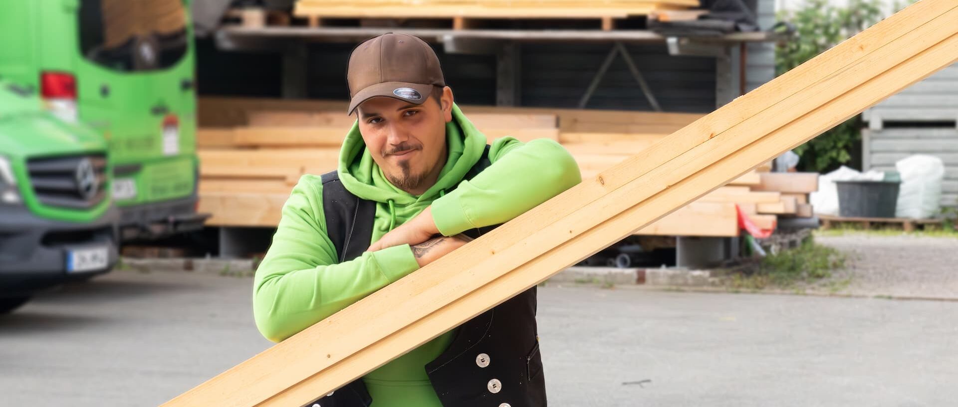 Christopher Porträtfoto Christopher - Er steht auf dem Firmengelände und stützt seine Arme auf mehreren Holzlatten ab, die schräg an einer Wand gestellt sind.